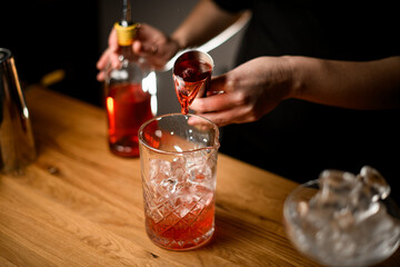 Female hands of a bartender pouring a jigger of brown liquid into a cocktail glass with ice