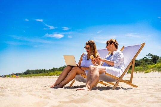 Remote work on beach. Two young women dressed in white sitting on deckchairs and working on laptop and smartphone on sandy beach on sunny day.