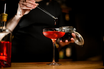 Focus on tall stemmed wine glass filled with brown cocktail above which female bartending hand holds berry