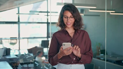 Young dreaming Indian woman manager using cell phone mobile AI app for work call, business communication, trading. Pondering Latin Hispanic female businesswoman holding smartphone standing in office - Powered by Adobe