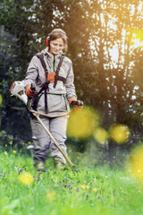 A woman in protective gear trims a green meadow with a gardening trimmer under the sun, engaging in...