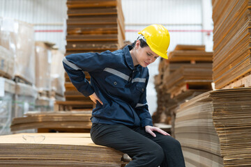 Female warehouse worker in a yellow helmet and blue uniform experiences back pain while sitting on stacked cardboard sheets at warehouse factory.