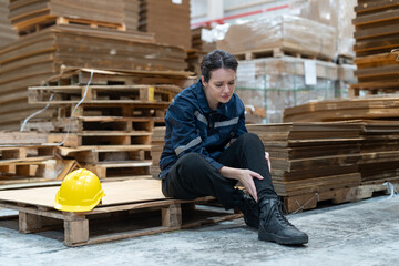 Female warehouse worker sits on a pallet in a warehouse, holding her injured leg with a concerned expression after working and checking stock products on shelves at warehouse factory.