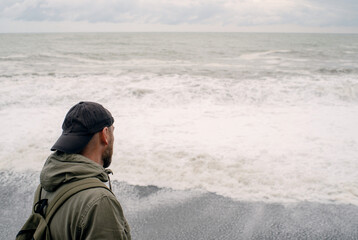 A lonely sad man in a cap and with a backpack looks into the distance at the stormy sea on a cloudy...