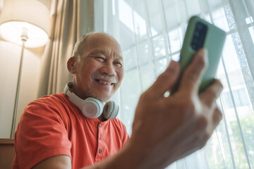 Asian senior man with glasses and headphones around neck smiling while using smartphone near window with curtains, video call with family