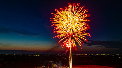 Large Red And Gold Fireworks Exploding In The Night Sky, Creating Brilliant Bursts Over The Distant City Lights And A Gathered Crowd, With A Vibrant Twilight Horizon Enhancing The Festive Atmosphere.