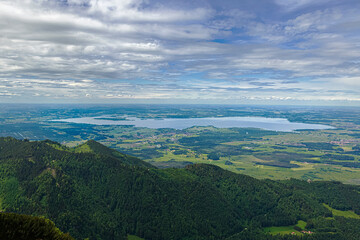 Obraz premium Weite Landschaft mit dem Chiemsee im Hintergrund, umgeben von grünen Hügeln und Bergen unter einem bewölkten Himmel, Chiemgau, Bayern, Deutschland