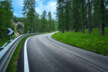 Fototapeta premium Road in green forest in summer. Dolomites, Italy. Beautiful mountain road, trees with lush foliage, grass, high rocks, blue sky with clouds. Landscape with empty curvy road in the woods. Transport