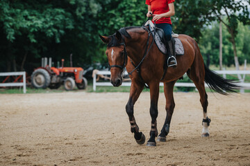 A girl wearing a red shirt rides a horse on a ranch, with a tractor visible in the background. Equestrian activity.