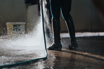 Equestrian washing horse legs with hose in stable yard after riding session, wearing riding boots...