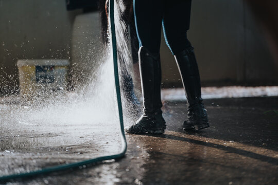 Equestrian washing horse legs with hose in stable yard after riding session, wearing riding boots and wetting the ground.