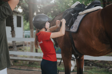 Young girl securing a saddle on a horse outdoors in a stable. Preparing for horse riding and equestrian activities. © qunica.com