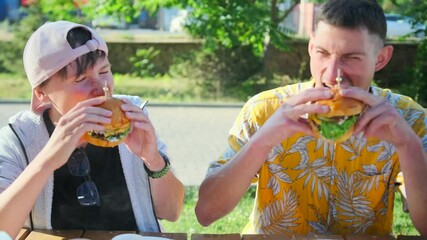 Two friends greedily bite juicy cheeseburgers smiling in a street cafe sitting at a table under the rays of the sun. Slow motion.