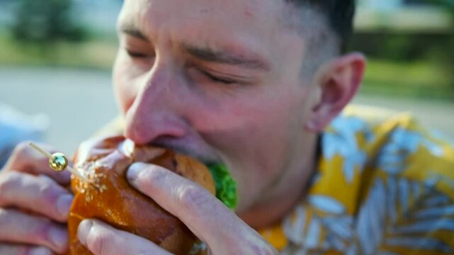 Close up, hungry young man bits burger while sitting with friends in a cafe outdoors. Slow motion.