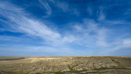 A bright blue sky with wispy white clouds above a dry badlands landscape.
