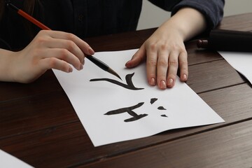Naklejka premium Calligraphy. Woman with brush writing words River and Entrance in Chinese on paper at wooden table, closeup