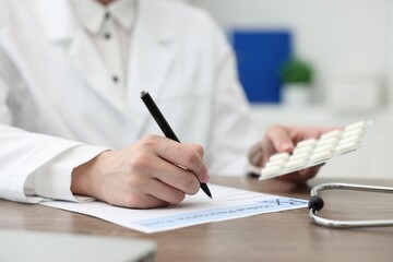 Doctor with pills writing prescription at wooden table in clinic, closeup