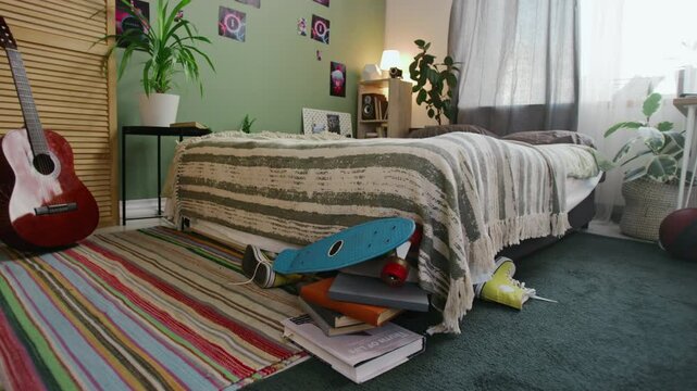 Full shot of pile of schoolbooks, skateboard, pair of sneakers lying on floor by bed, guitar standing against wardrobe, houseplants and posters in young male student or teenage boys bedroom