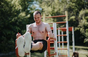 A fit man exercises on outdoor fitness equipment in a park, focusing on strength training. He is...
