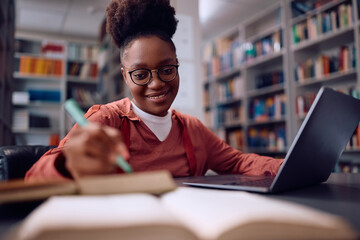 Happy black female student e-learning on laptop in library.