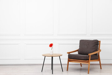 Comfortable armchair and coffee table with gerbera flower near white wall, space for text. Interior design