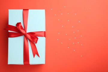 Gift box with red bow and confetti on coral background, top view. Space for text