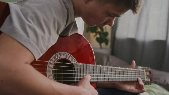 Medium close-up tilt shot of young man with hearing impairment, wearing cochlear implant device behind ear, sitting in bedroom, strumming guitar strings, while training sound perception skills
