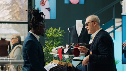 Experienced famous stylist buying the perfect tie color for his rich client, looks for expensive materials at shopping center. Senior fashion designer giving orders to store woman employee. Camera A.