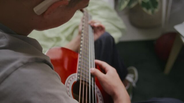 Medium over-shoulder shot of unrecognizable young man with electronic hearing aid sitting on bed at home and playing acoustic guitar to improve sound perception capacity