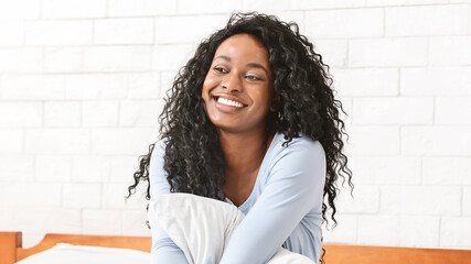 Young african cheerful woman sitting on bed and smiling, hugging pillow, free space