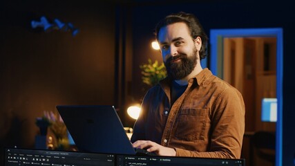 Portrait of smiling man doing IT support job from home, standing in personal office with laptop in...