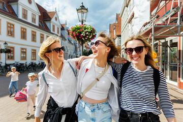 Three cheerful young adult women friend with kids walk street old european german town city street...
