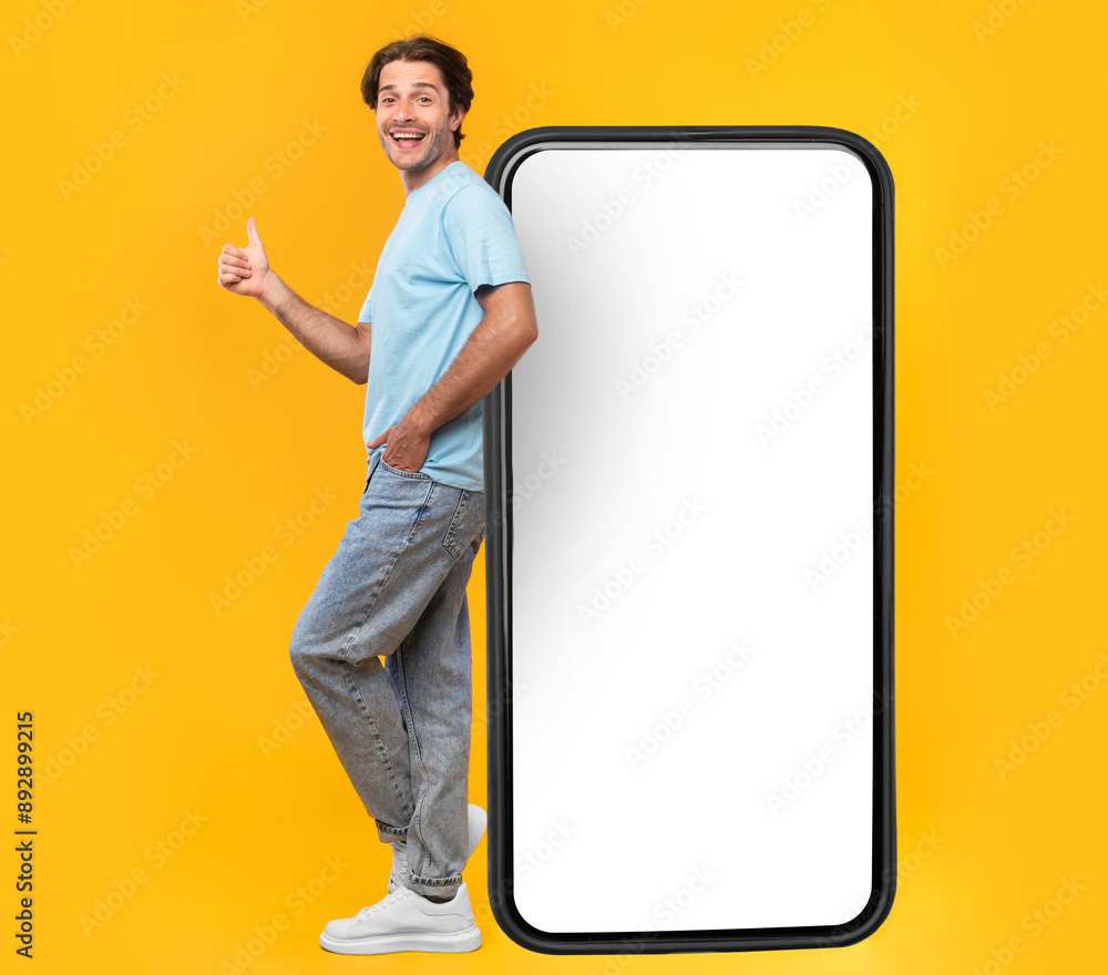 Poster excited man leaning on big smartphone with blank white screen and gesturing thumb up sign, cheerful 