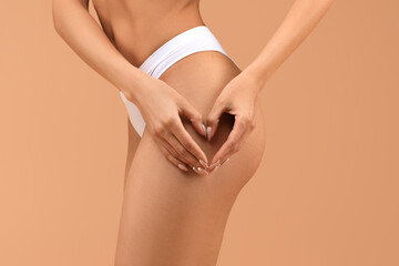 Cropped of woman in white underwear stands with her back turned to the camera, using both hands to form a heart shape over her left thigh. Her skin is smooth and the background is a soft peach color.