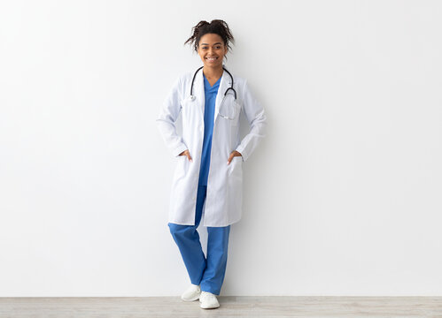 Friendly female black doctor posing with hands in pockets looking at camera standing leaning on wall, lady wearing blue uniform, coat and stethoscope, full body length, white studio background banner
