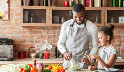 Black smiling girl adding olive oil to veggies salad, dad mixing vegetables in bowl, kitchen interior, copy space