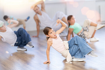 Smiling sports teen girl performing krump movements on floor with group children in modern breakdance studio with male coach