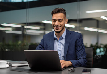 Fototapeta premium Smiling young middle eastern businessman sitting at worktable at modern office, typing on computer keyboard, sending emails to his business partners, working on marketing research, copy space