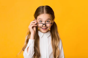 Portrait Of Cute Schoolgirl Looking At Camera Over Eyeglasses On Yellow Studio Background.