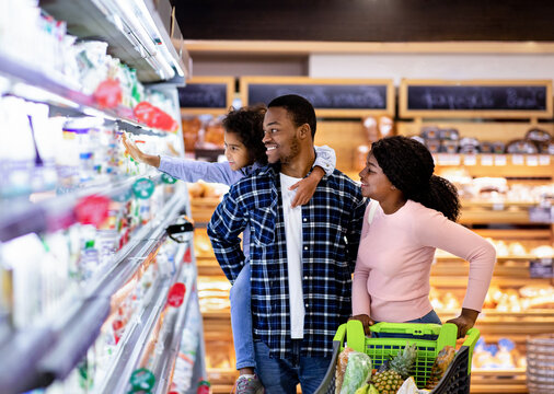 Portrait of happy black family with trolley shopping together at grocery store. Millennial African American parents with lovely daughter picking food, choosing milk products at big mall