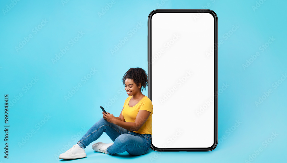 Poster cheery african american lady sitting near big cellphone with empty white screen, using mobile device