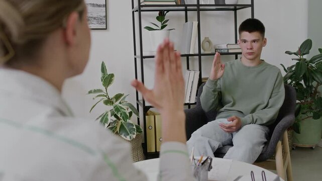 Medium over-shoulder shot of teenage Caucasian boy with hearing impairment sitting in classroom, reciting American sign language alphabet with female teachers help