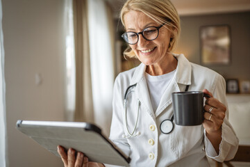 Portrait of mature woman doctor stand and hold cup and digital tablet