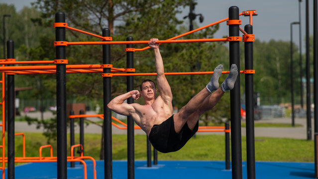 Young muscular man hanging by one hand on a horizontal bar outdoors. 