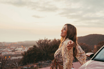 Young beautiful woman enjoying summer and having fun sitting on the hood of the car opening arms feeling good and free. Freedom concept and happy lifestyle at sunset.