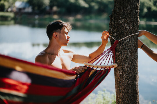 Young man setting up a colorful hammock by a tree near a serene lakeside on a sunny day, enjoying nature and relaxation.