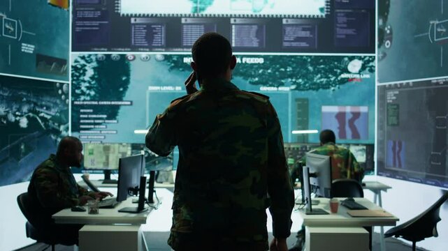Military man answers phone call in a mission control center, working with advanced technology and satellite imagery to track potential threats and real time data. Intelligence activities. Camera B.