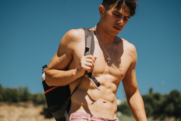 Shirtless young man with a muscular physique and backpack enjoying a sunny day outdoors near the...