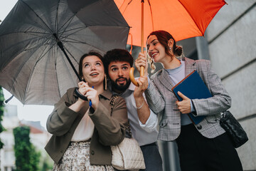 Young business colleagues share umbrellas during a rainy outdoor meeting in the city. They...