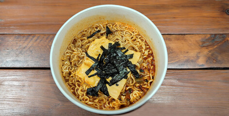 korean instant noodles with spicy sauce topped with cheese and seaweed slices served in a paper bowl, served warm isolated on a brown wooden table